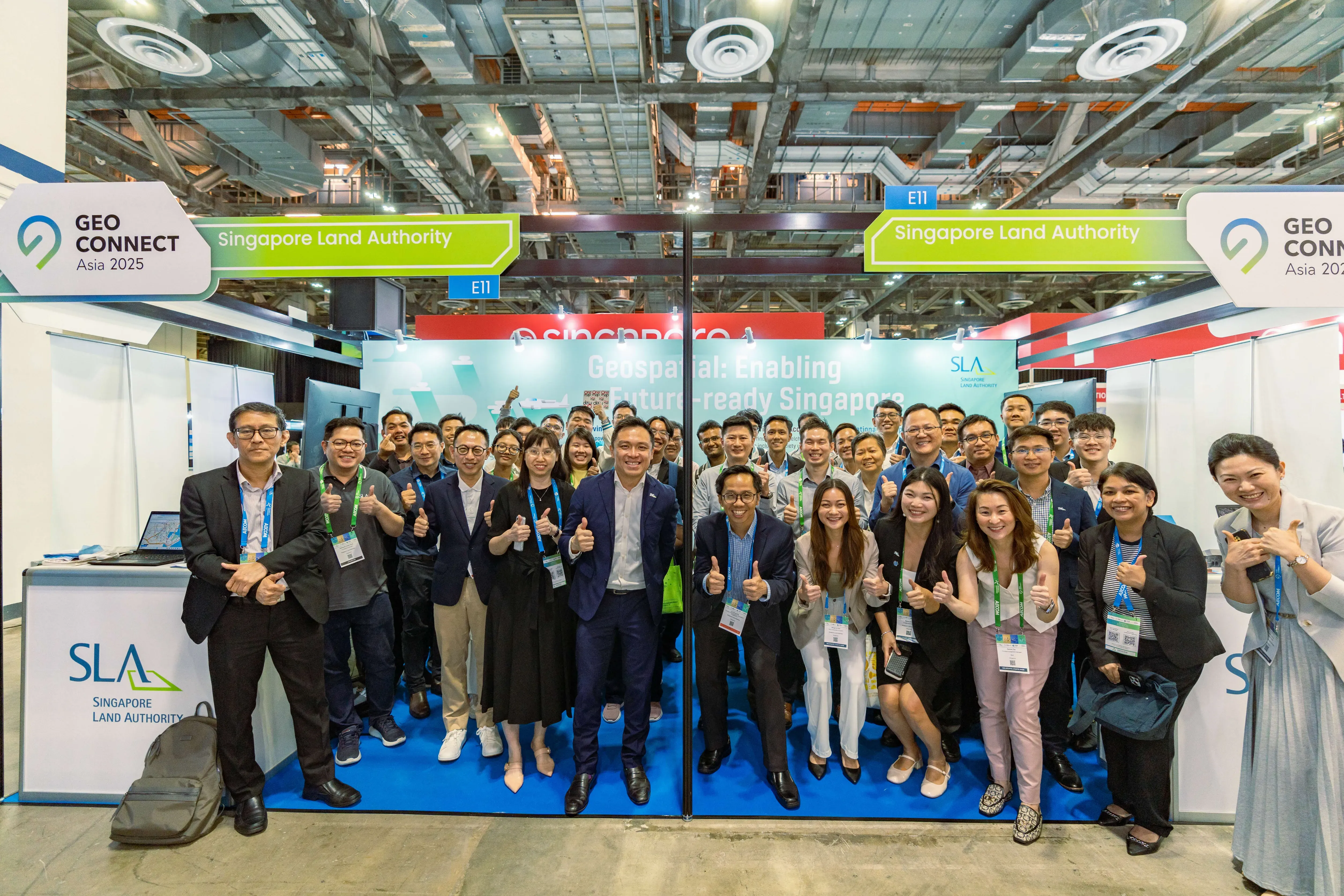 Large group posing with thumbs up at Singapore Land Authority booth E11 at Geo Connect Asia 2025 expo.